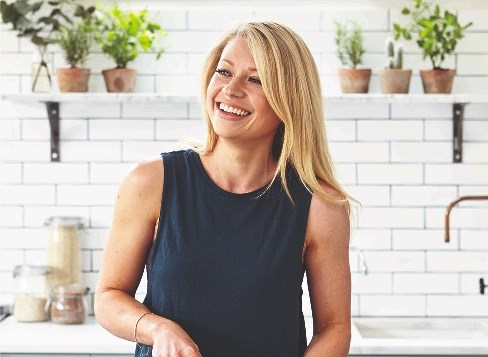 Hollie Grant laughs in a white-tiled kitchen with plants on the walls.