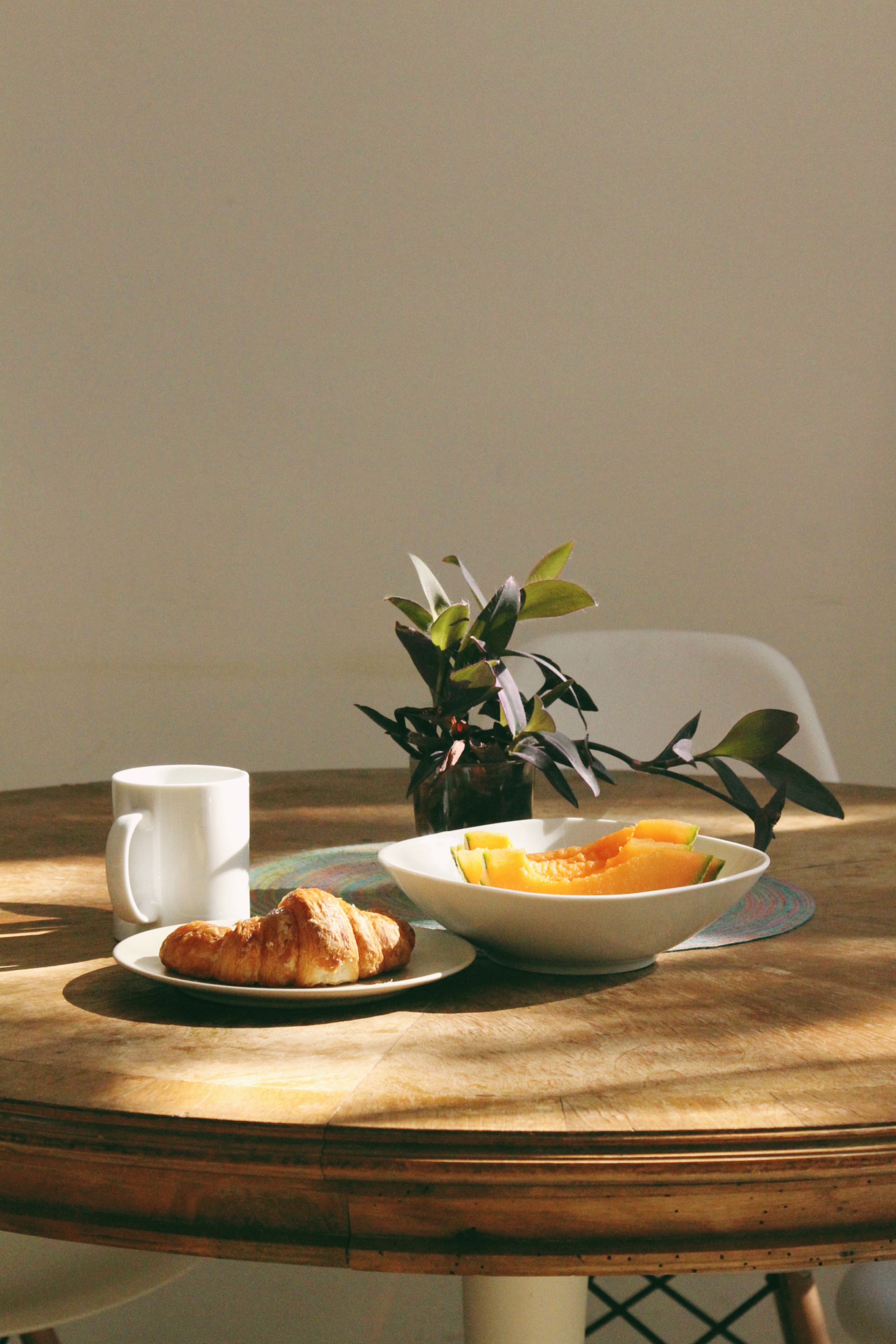 A breakfast sits of a white mug of coffee, a croissant, and two slice of cantaloupe sit on a wooden table alit in morning sunshine with a green plant as centrepiece. Photo by Tatiana from Pexels.