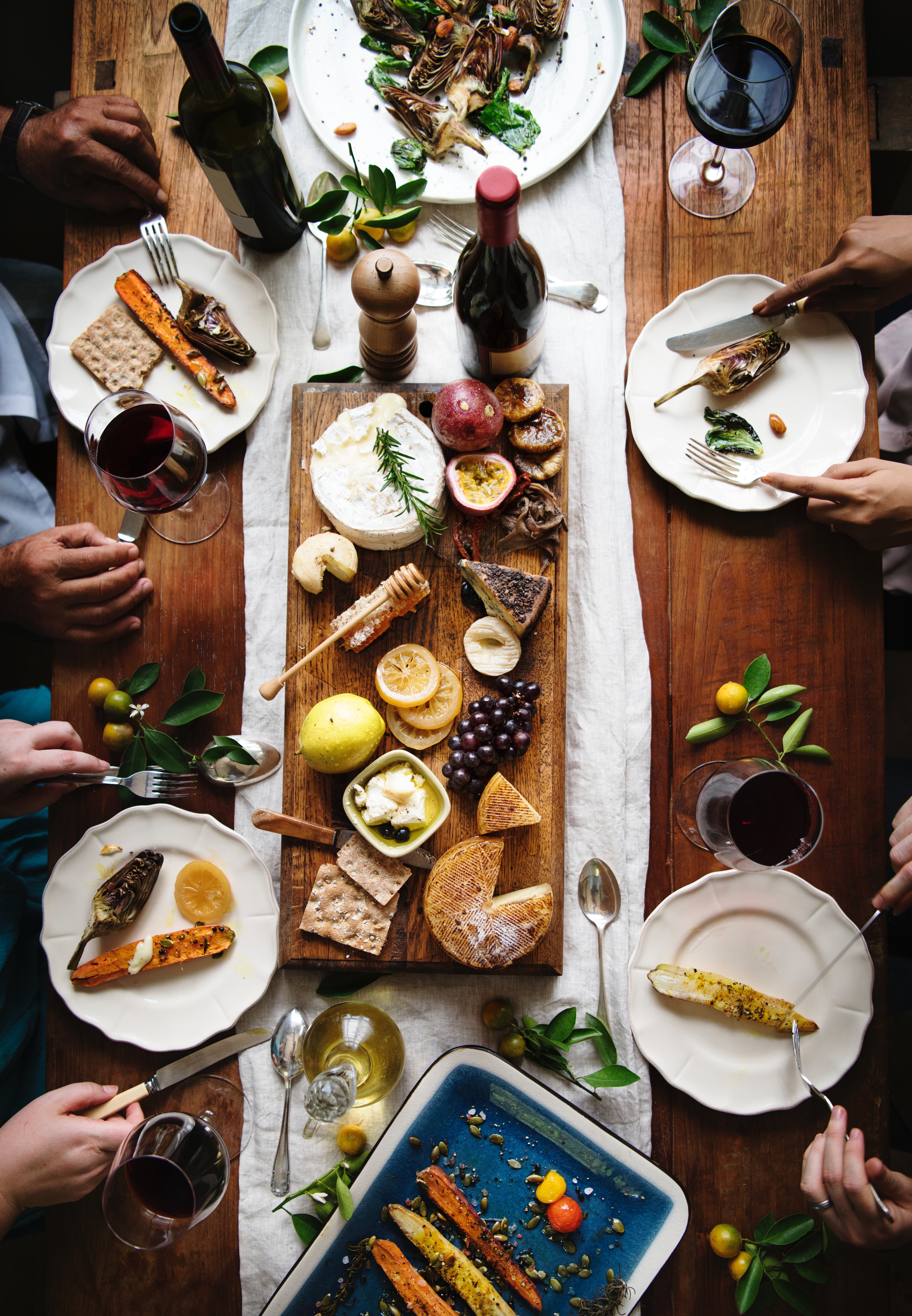 Shot from above, a group of four people share a plate of cheeses, fruit, grilled veggies, and red wine. Photo by rawpixel.com from Pexels.