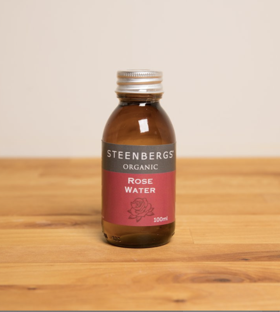 A 100ml brown bottle with a red, grey, and white label reading 'Steenbergs' Organic Rose Water' is contained by a silver twist cap. The bottle sits on a light-coloured wooden table with a grey wall behind it. 