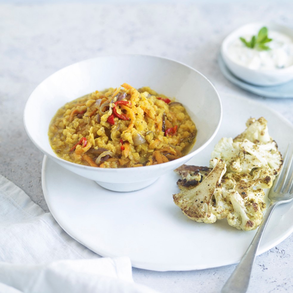 A white bowl of Lentil Dal + spiced roast cauliflower sits on a whiter table. A side of roast cauliflower and a bowl of tzatziki are present in the background.