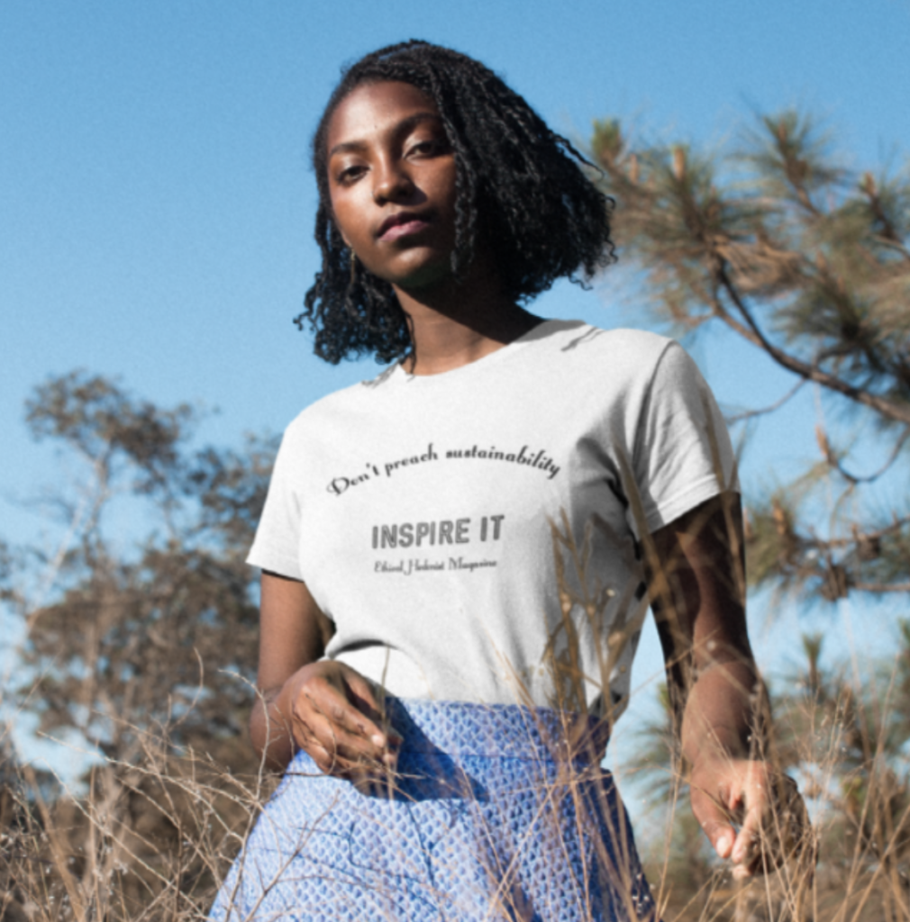 A model wears the fitted 'Don't Preach Sustainability, Inspire It' t-shirt made in collaboration between Teemill and Ethical Hedonist. She is outdoors in front of tall trees with tall grass in front of her.