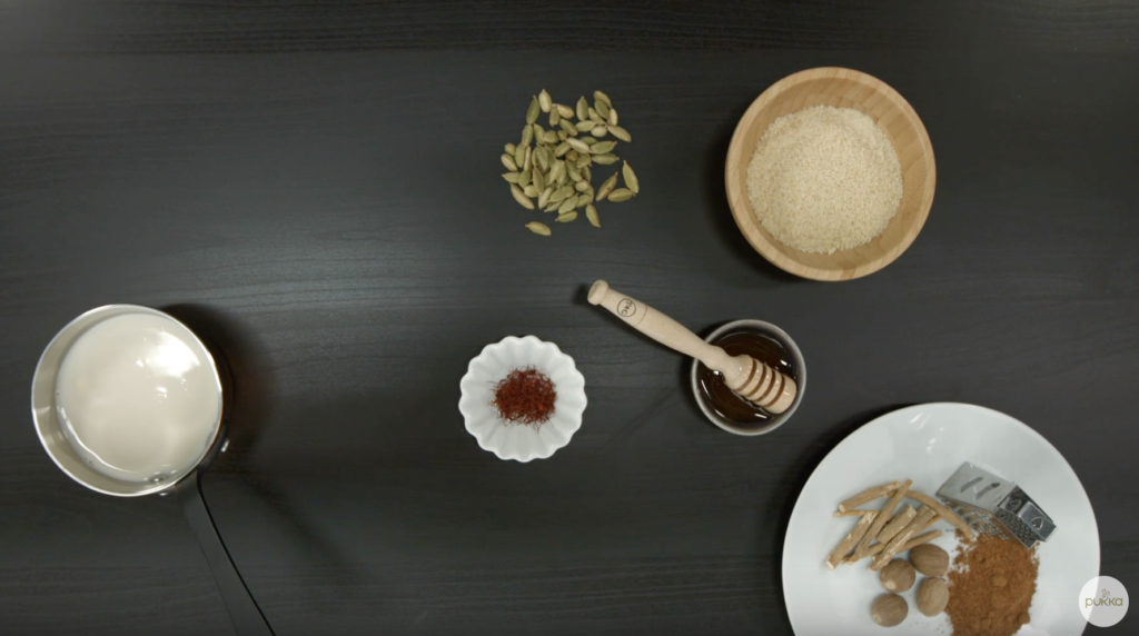 A flat-lay of all the ingredients required for the Golden Milk of Bliss recipe, including cardamom pods, crushed almonds, honey, saffron, ashwagandha root, nutmeg, turmeric root powder, and almond milk.