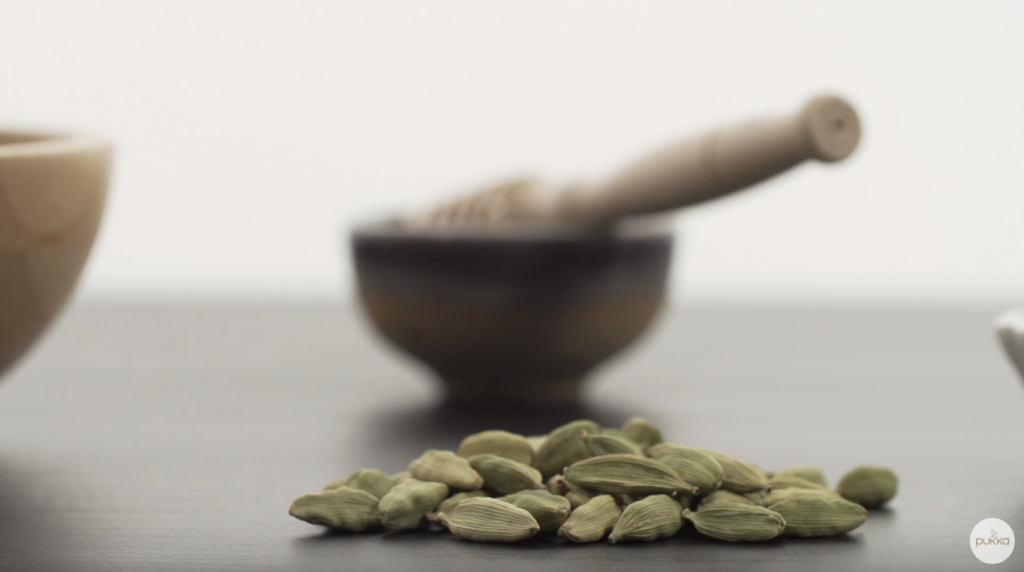 A close-up of cardamom pods with a blurred bowl of honey in the background.