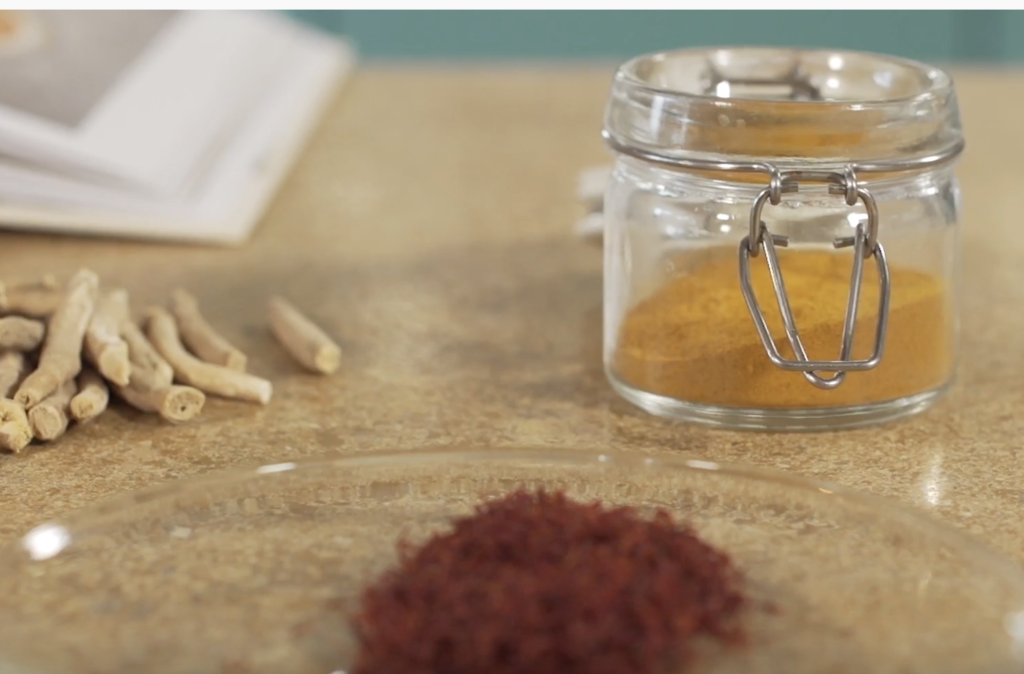 A container of turmeric powder sits in the background while piles of saffron and ashwagandha root sit blurred in the foreground.