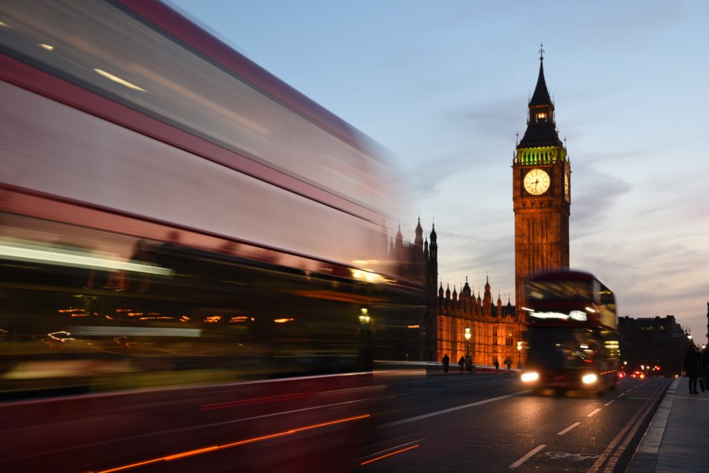 Two blurry double-decker buses pass by the Houses of Parliament in Westminster, London at dusk.